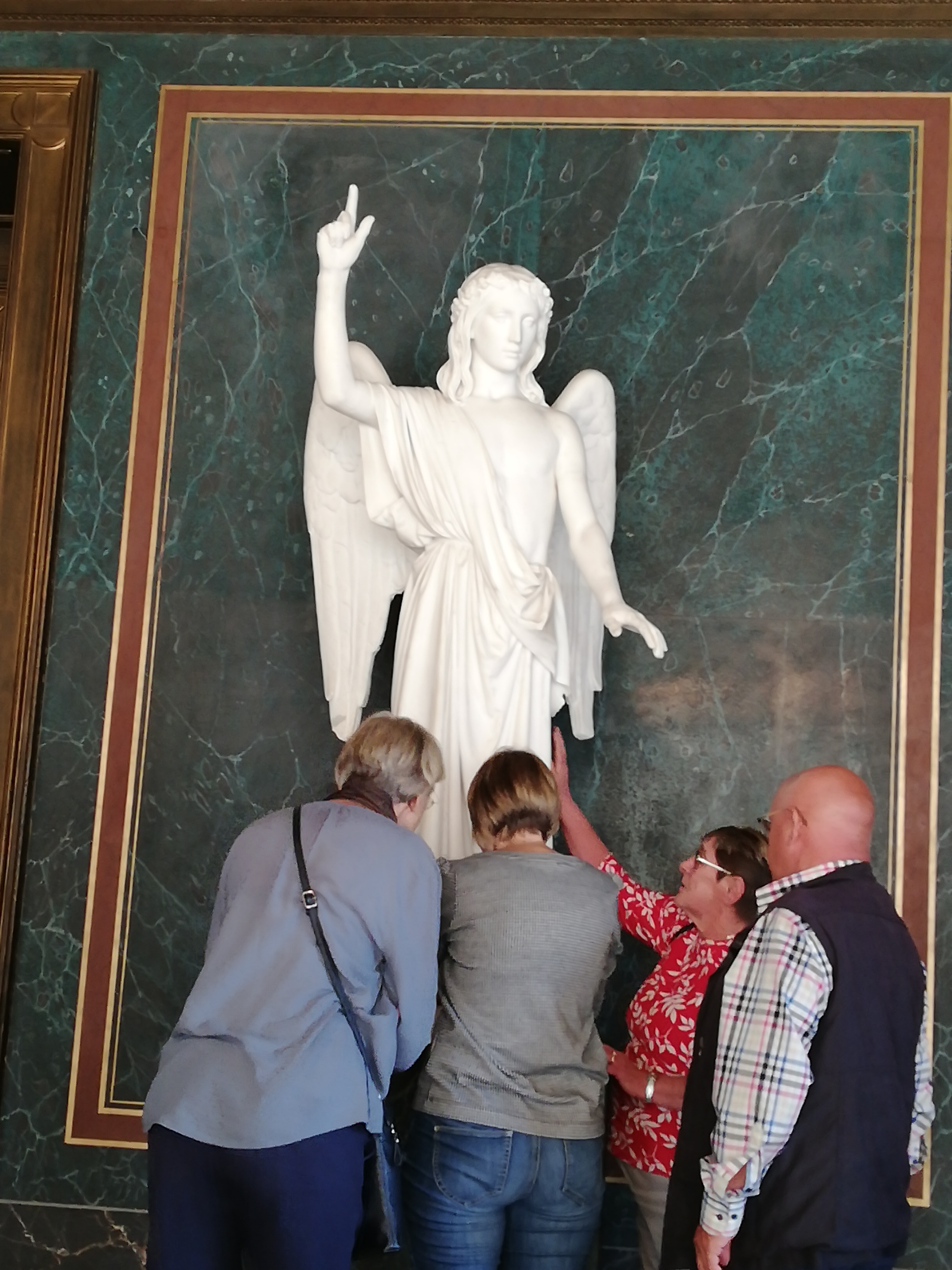 Vor einem großen weißen Engel stehen vier Menschen. Drei von ihnen ertasten die Figur.